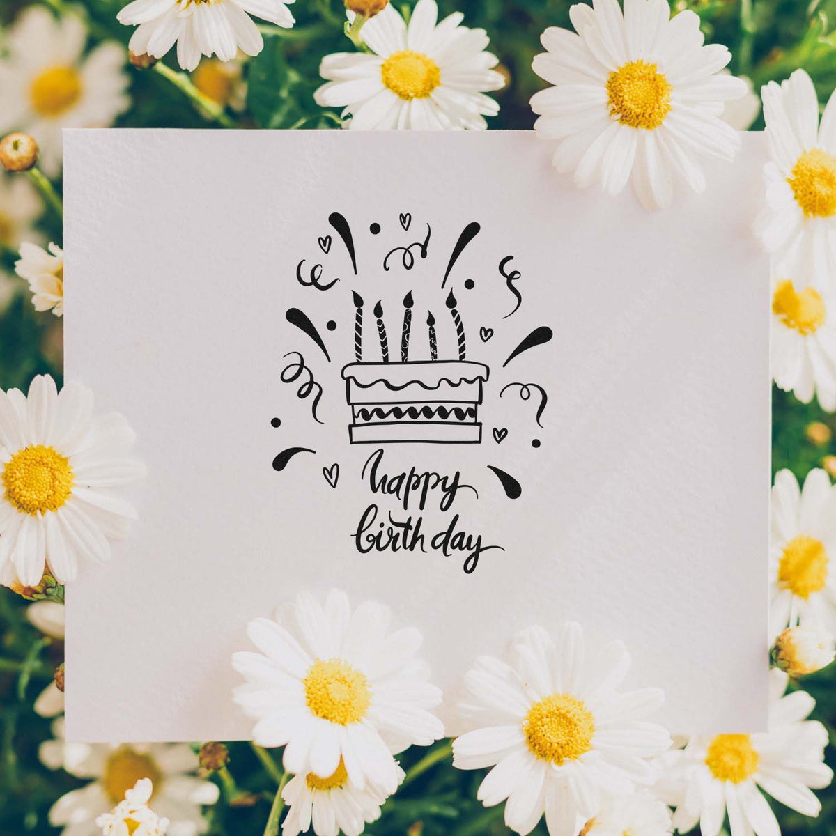 Black happy birthday cake stamp on a white card, surrounded by white daisies with yellow centers.