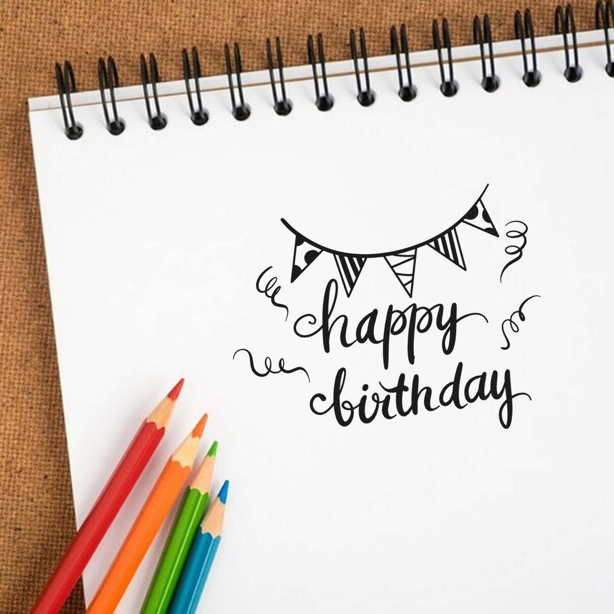 Black happy birthday stamp seal on white spiral notebook, with bunting and colorful pencils on a brown background.