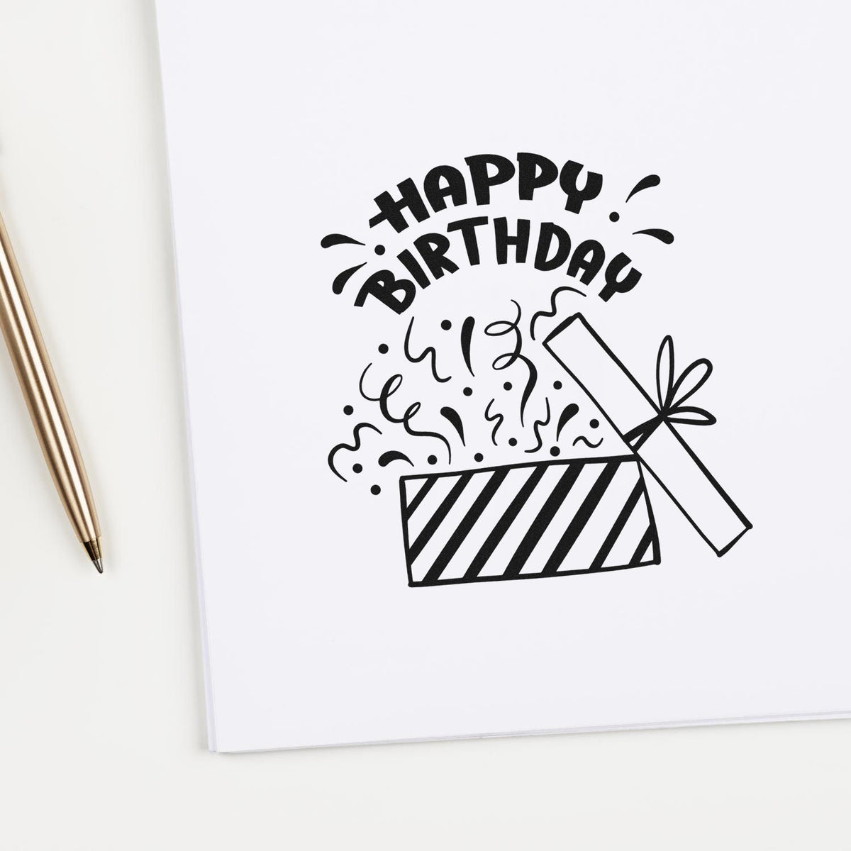 Black Happy Birthday stamp and seal design with confetti and striped gift box on white paper, beside a gold pen