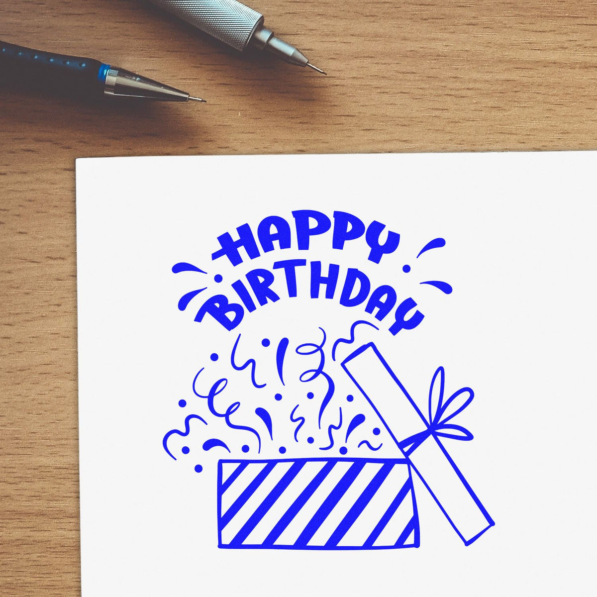 Blue Happy Birthday stamp and seal design with confetti, gift box, and two pens on a wooden desk.