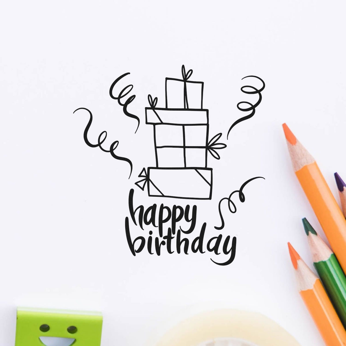 Black happy birthday stamp seal with stacked gift boxes and confetti, shown on white paper beside orange and green pencils.