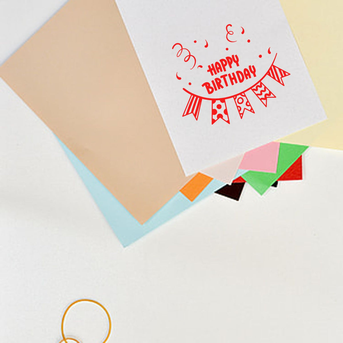 Red Happy Birthday stamp on white card with colorful bunting, layered pastel papers, and seal-style design samples.