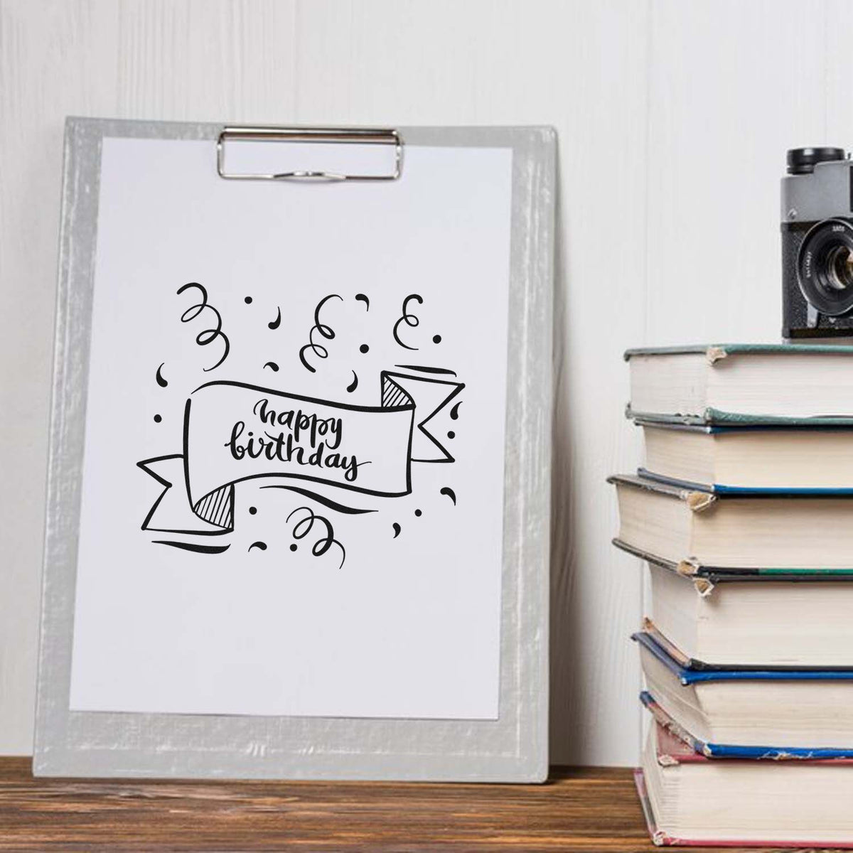 Black happy birthday stamp design on white paper clipped to a gray clipboard beside stacked books.
