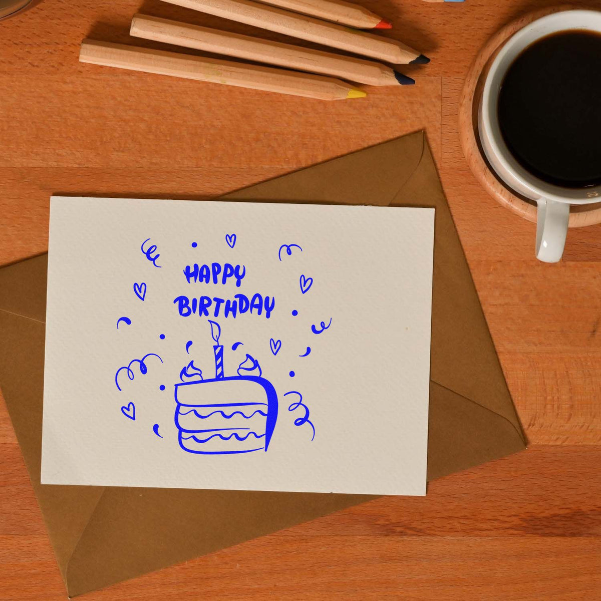 Blue birthday cake stamp and seal on white card, with brown envelope, pencils, and coffee on a wooden desk.