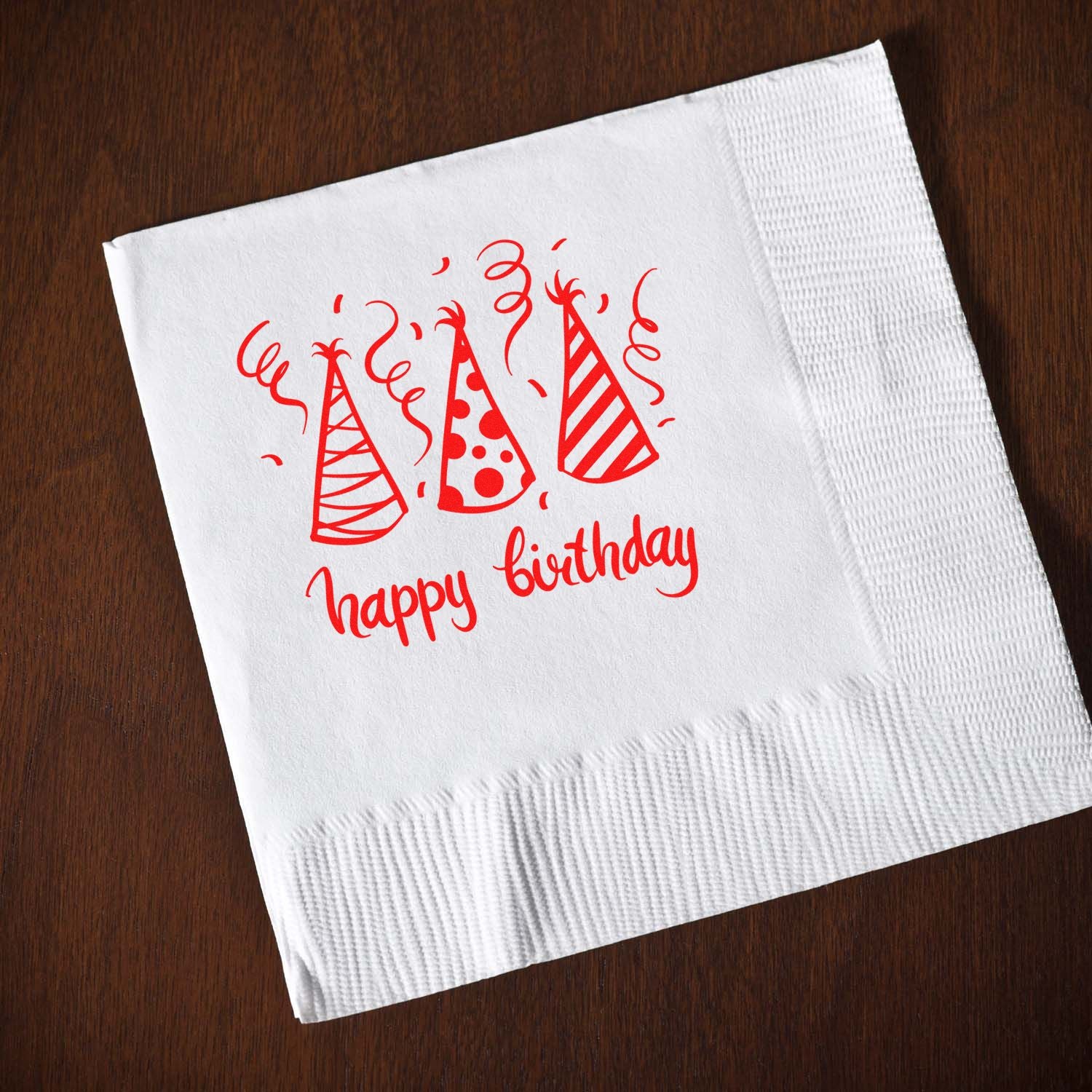 Red birthday stamp and seal design on a white napkin, with party hats and confetti on a dark wood background.