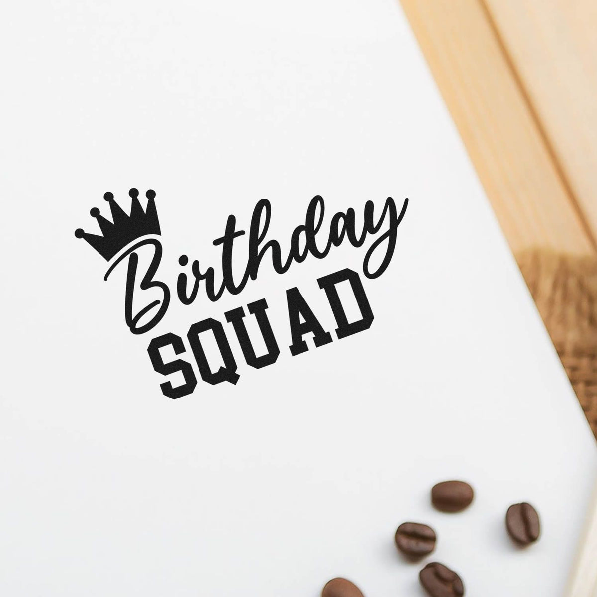 Black Birthday Squad stamp seal with crown on white paper, accented by coffee beans and a wood edge.