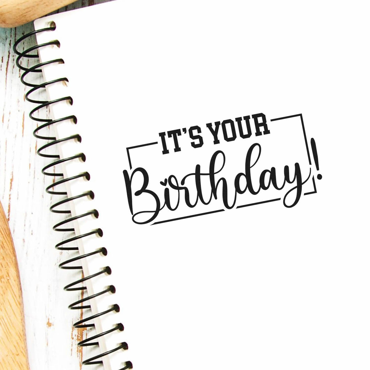 Black Its Your Birthday! stamp seal on a white spiral notebook, with rustic wood accents.
