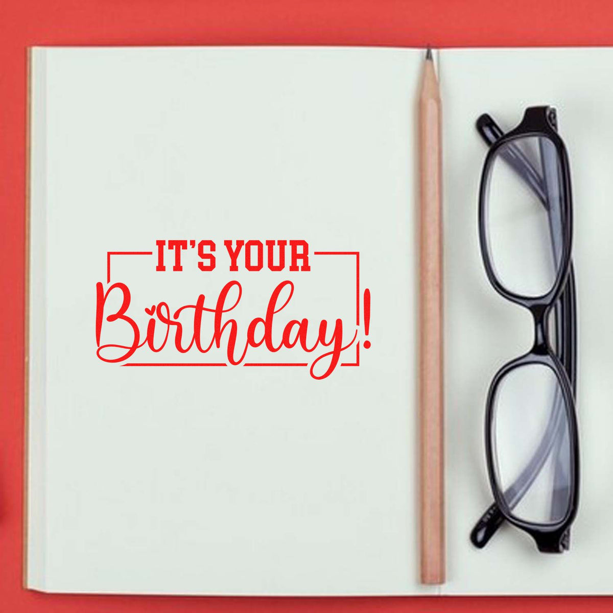 Red birthday stamp and seal design on open white notebook, with pencil and black eyeglasses.