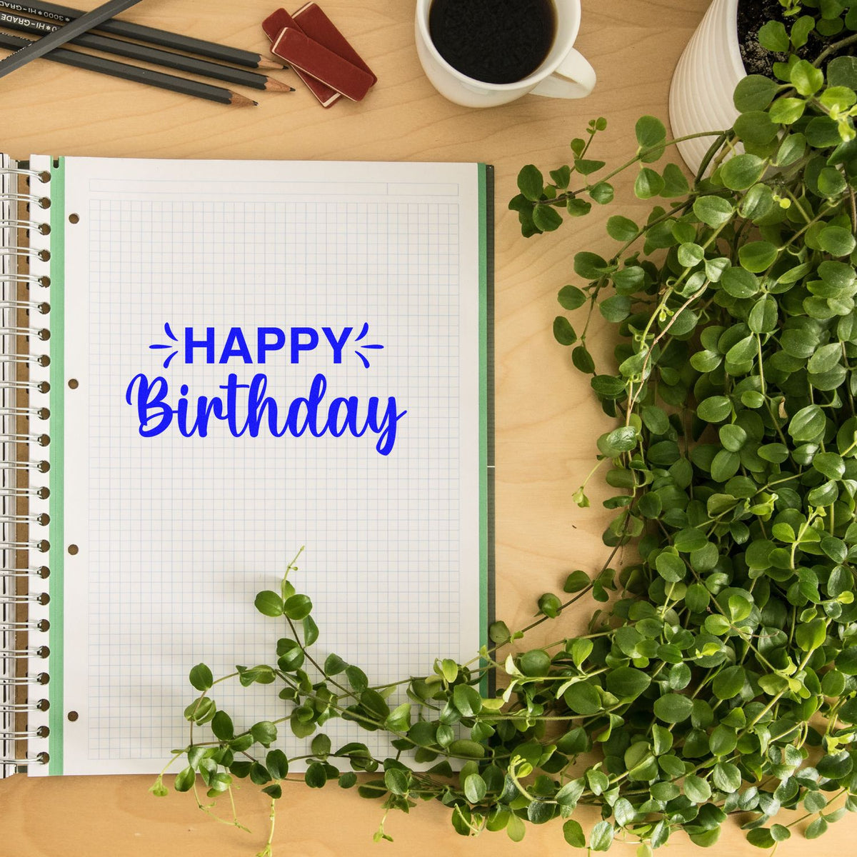 Blue Happy Birthday stamp on grid notebook with red seal blocks, pencils, coffee, and green plant on a wooden desk.