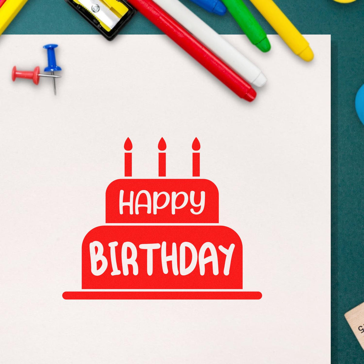 Red Happy Birthday cake stamp/seal on white paper, with colorful pens and push pins on a teal background