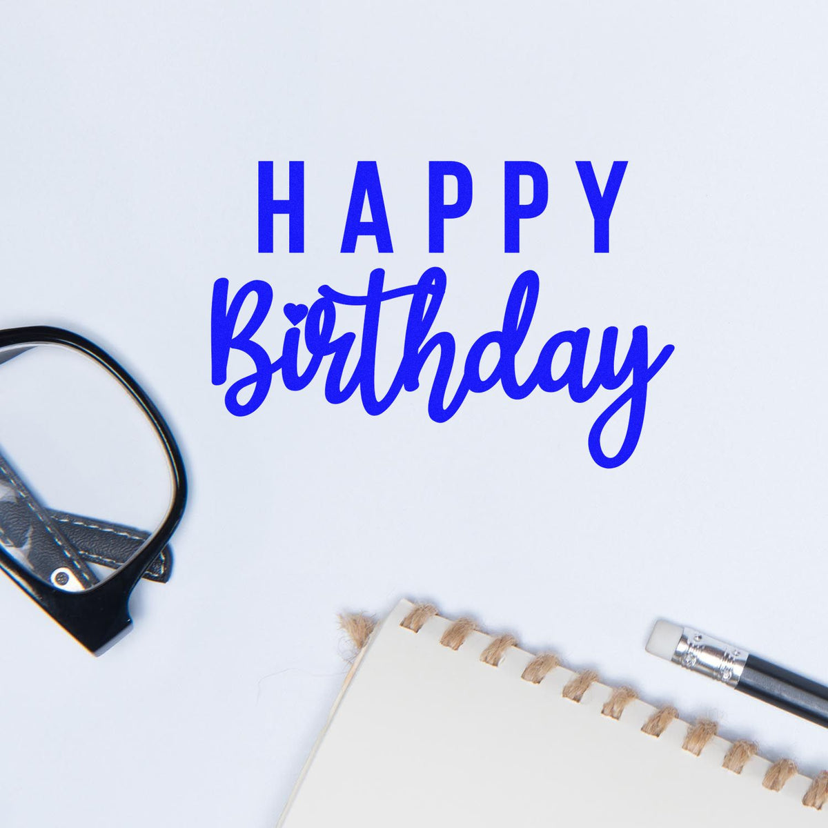 Blue Happy Birthday lettering with black stamp seal tool, notebook edge and pencil on white background