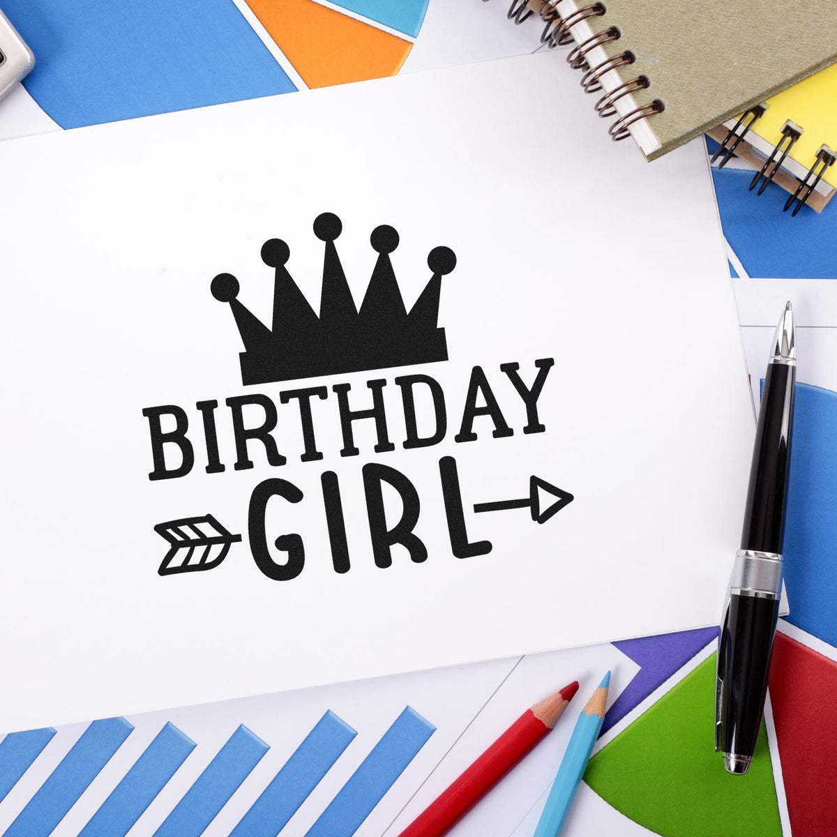 Black birthday girl stamp with crown on white paper, surrounded by colorful envelopes, notebooks, and pens.