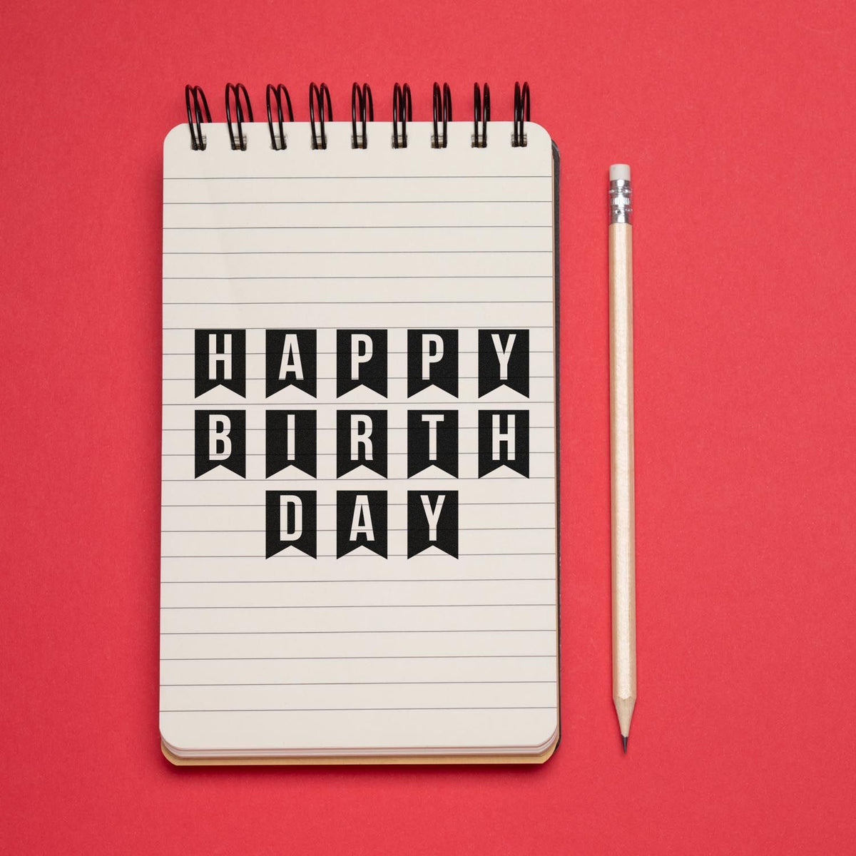 Happy Birthday stamp and seal design on a white spiral notebook, black banner letters, with pencil on a bright red background.
