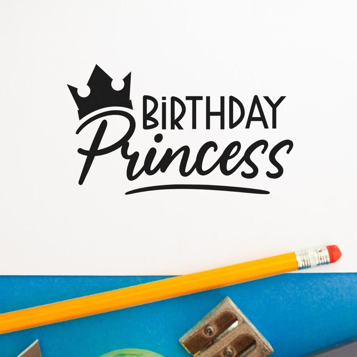 Black Birthday Princess stamp with crown on white paper, beside a yellow pencil on a blue surface.