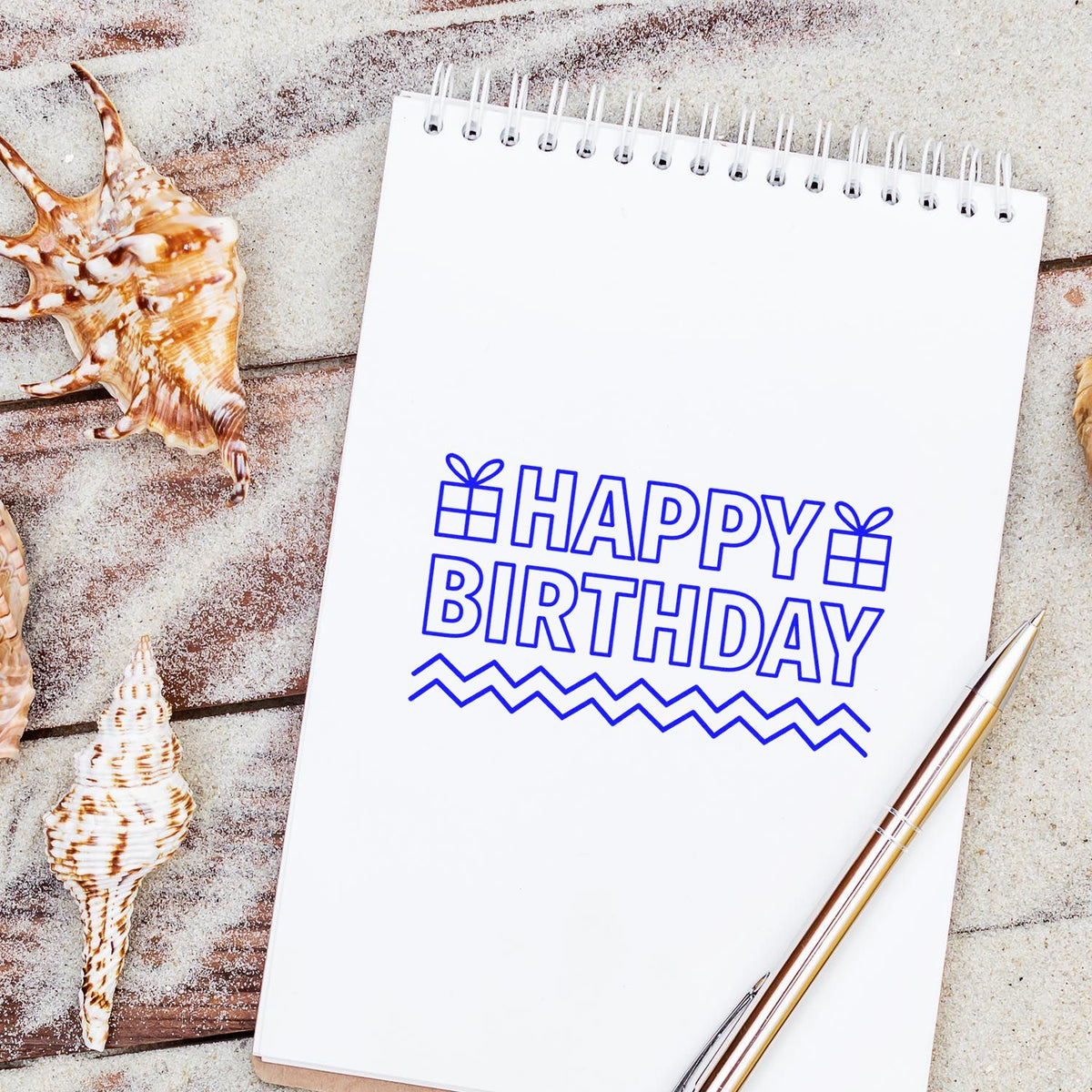 Blue Happy Birthday stamp and seal on a white spiral notepad, with gift icons, seashells, and a gold pen.