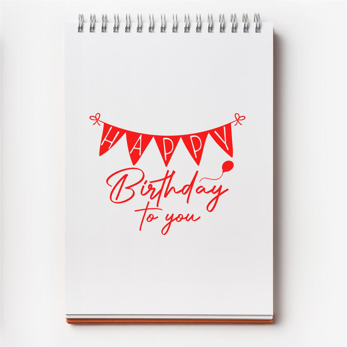 Red birthday stamp and seal design on white spiral notebook, with Happy Birthday bunting and balloon.