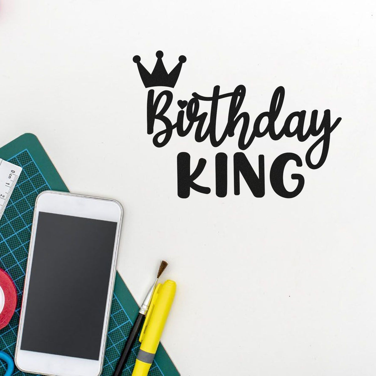 Black Birthday KING crown stamp/seal design on white, with green cutting mat, smartphone, yellow marker and craft tools.