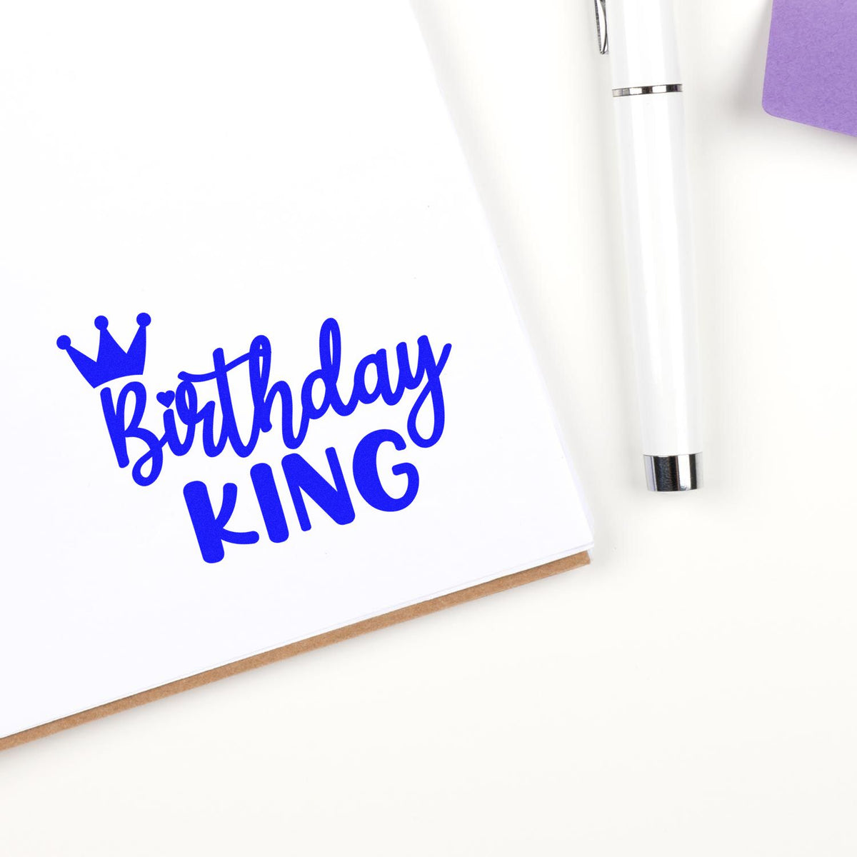 Blue Birthday KING crown stamp and seal on white card, beside a white pen with silver accents and a purple corner.