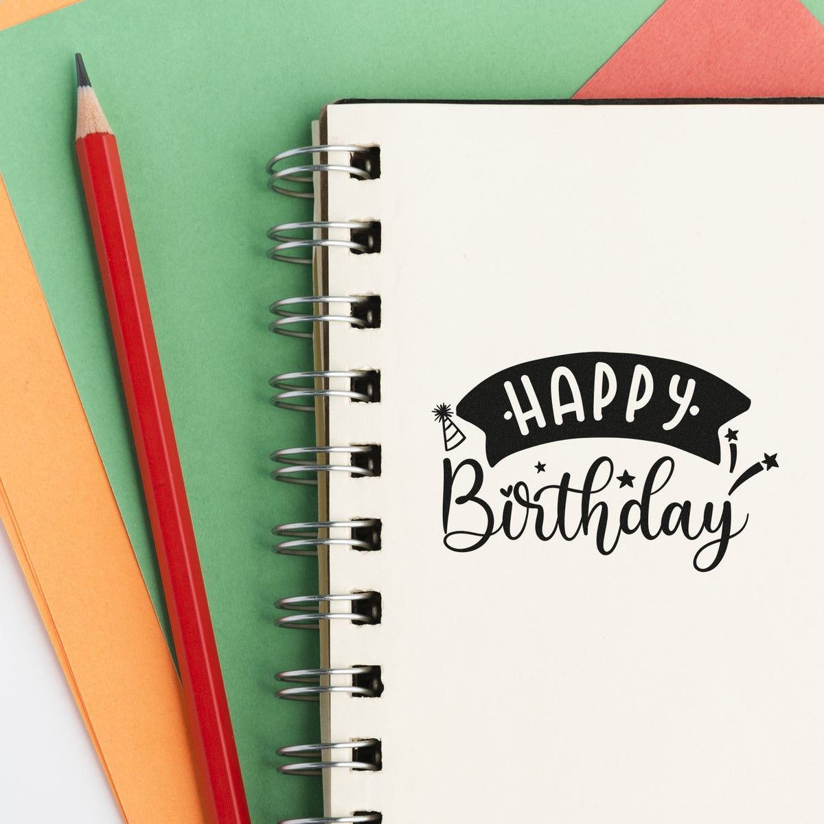 Black Happy Birthday stamp on a cream spiral notebook, beside a red pencil on green, orange and coral paper.