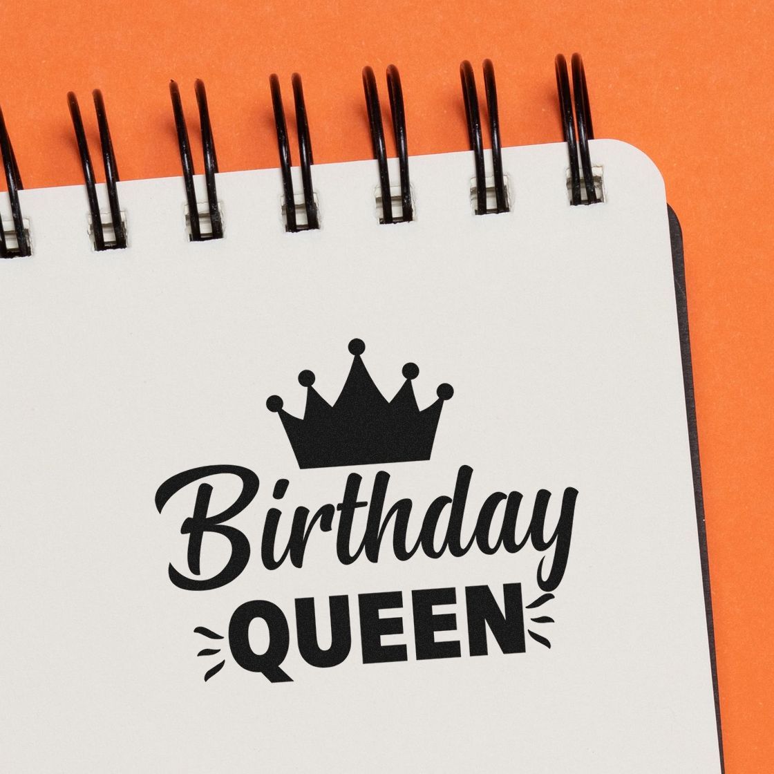 Black Birthday Queen stamp and seal design with crown on white spiral notebook against orange background.