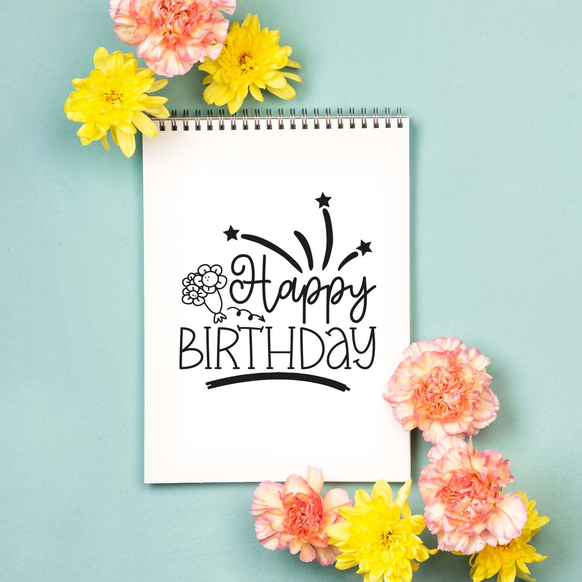 Black Happy Birthday stamp and seal on a white spiral notebook, framed by pink and yellow flowers on a mint green background.