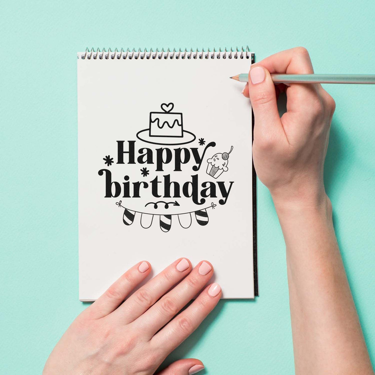 Black Happy Birthday stamp design with cake, cupcake and bunting on a white notebook, held by hand on a mint background.