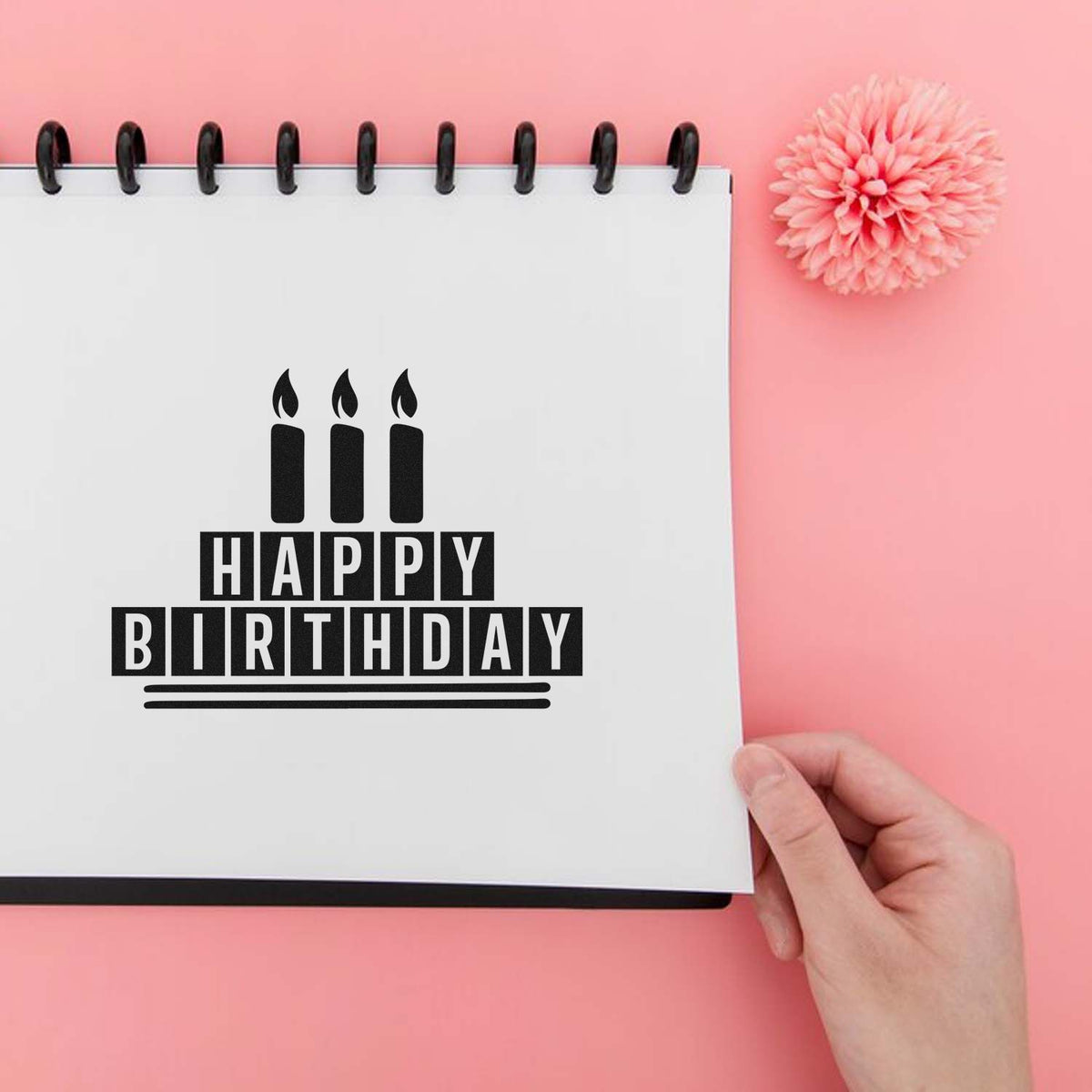 Black Happy Birthday stamp and seal on white spiral notebook, with candle graphic, pink background, and hand holding page.