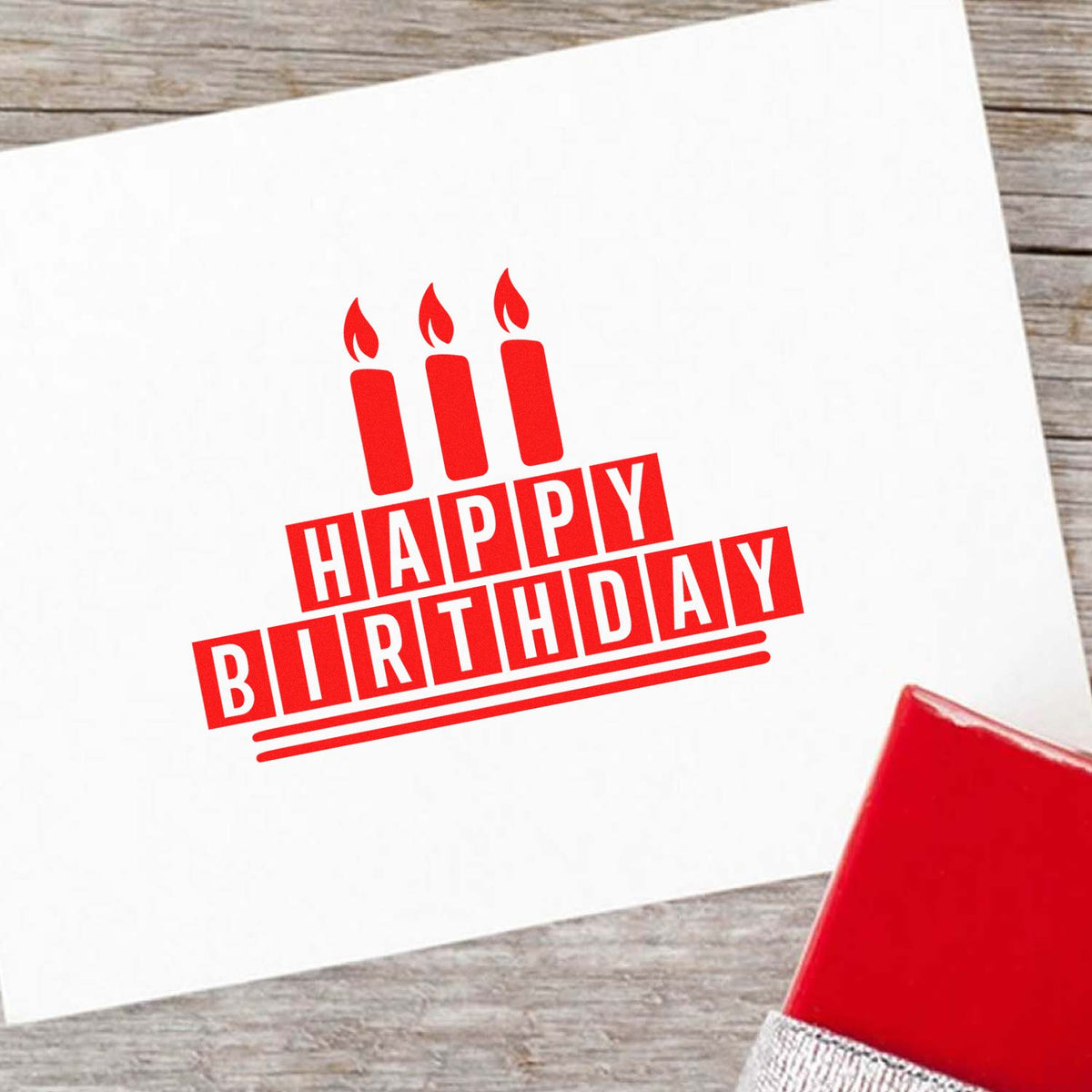 Red Happy Birthday rubber stamp with candles on white card, beside a red stamp pad on wood.