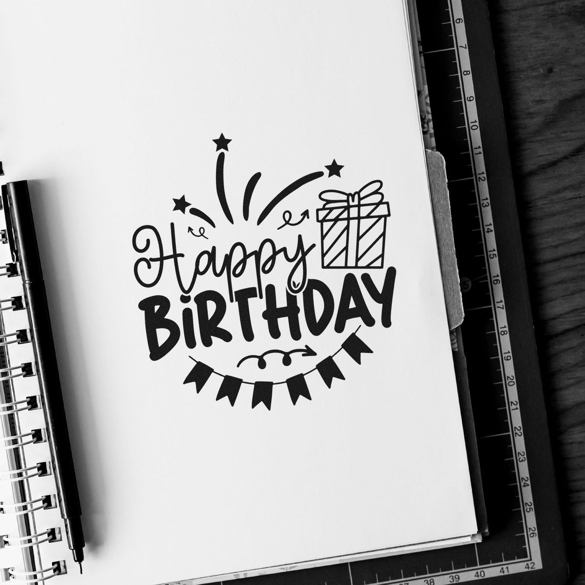 Black stamp and seal Happy Birthday design with gift, stars, and bunting on a white notebook page.