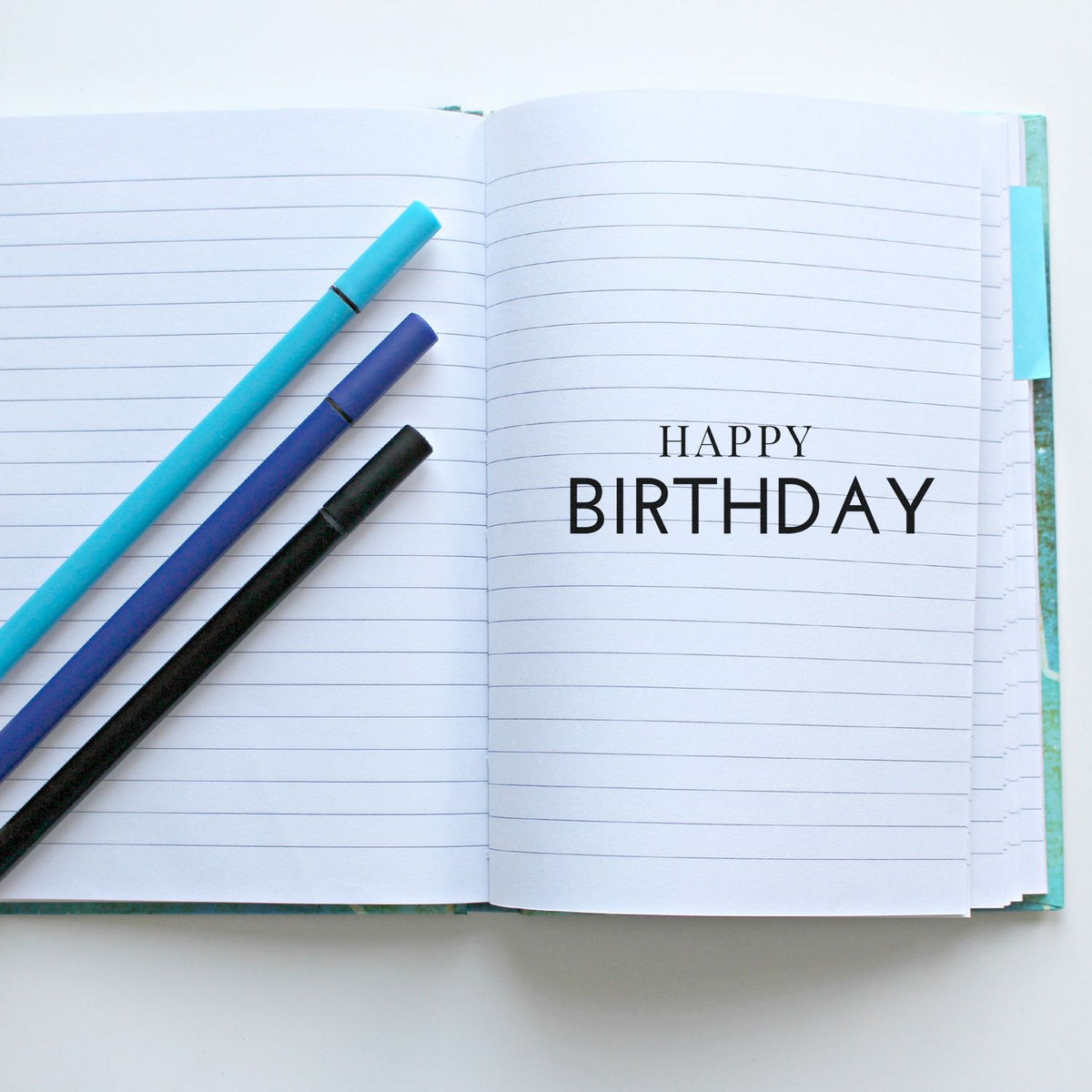 Open lined notebook with a black Happy Birthday stamp and three colorful stamp pens in turquoise, purple, and black.