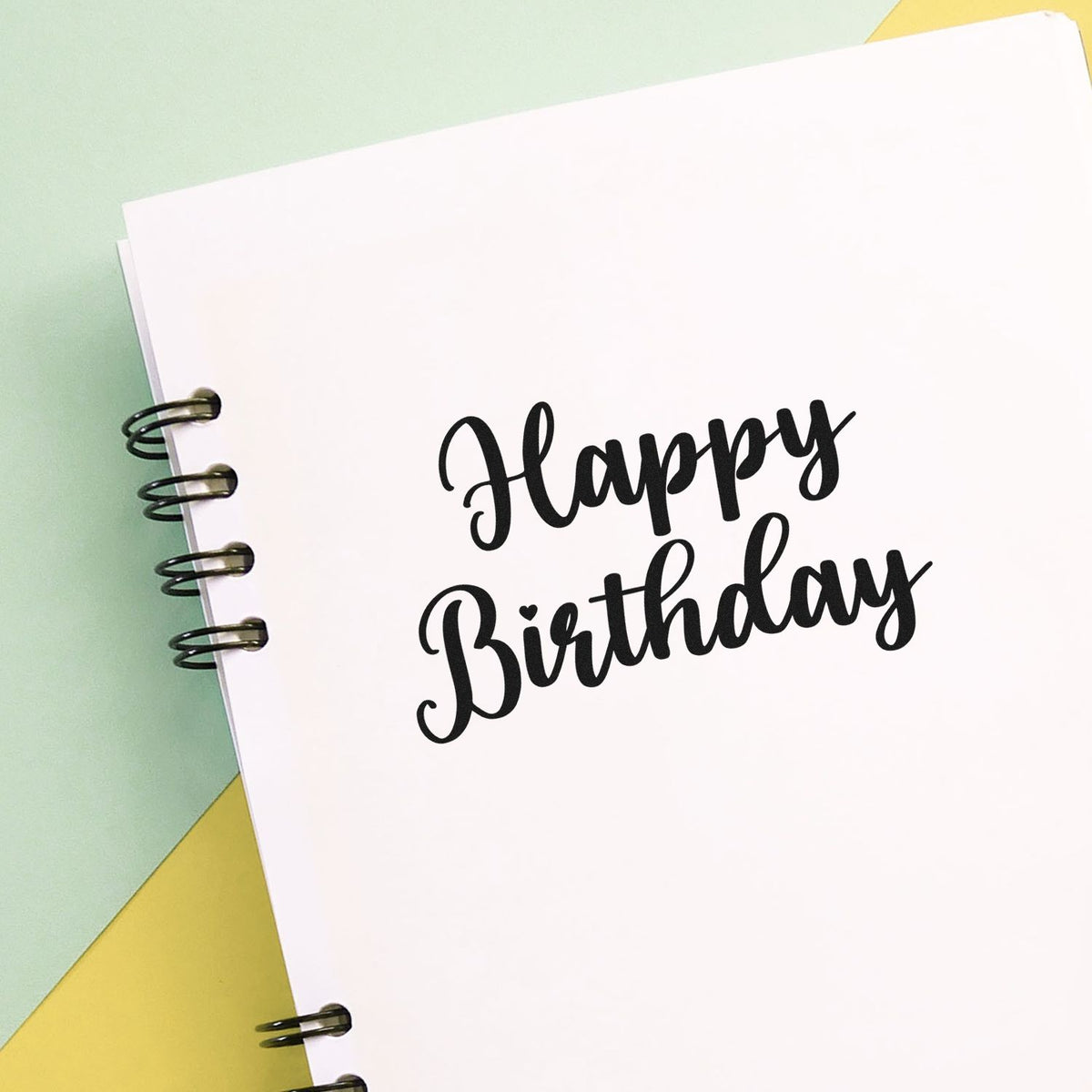Black Happy Birthday script stamp on white paper, with spiral binding, over mint green and yellow background.