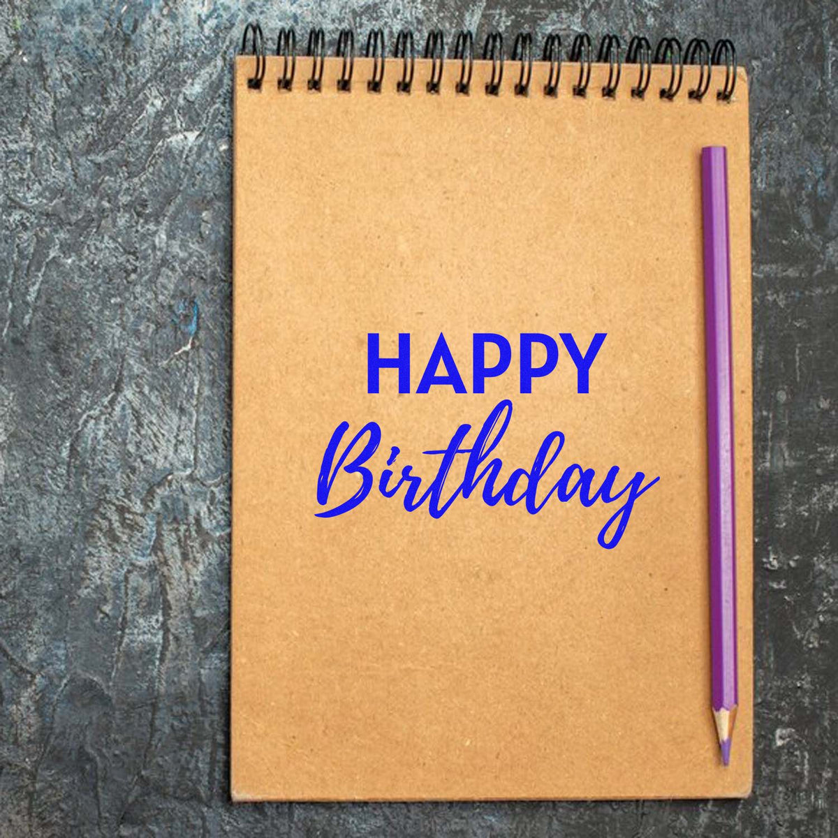 Happy Birthday stamp and seal on tan spiral notebook, bright blue lettering, with a purple pencil on dark textured background.