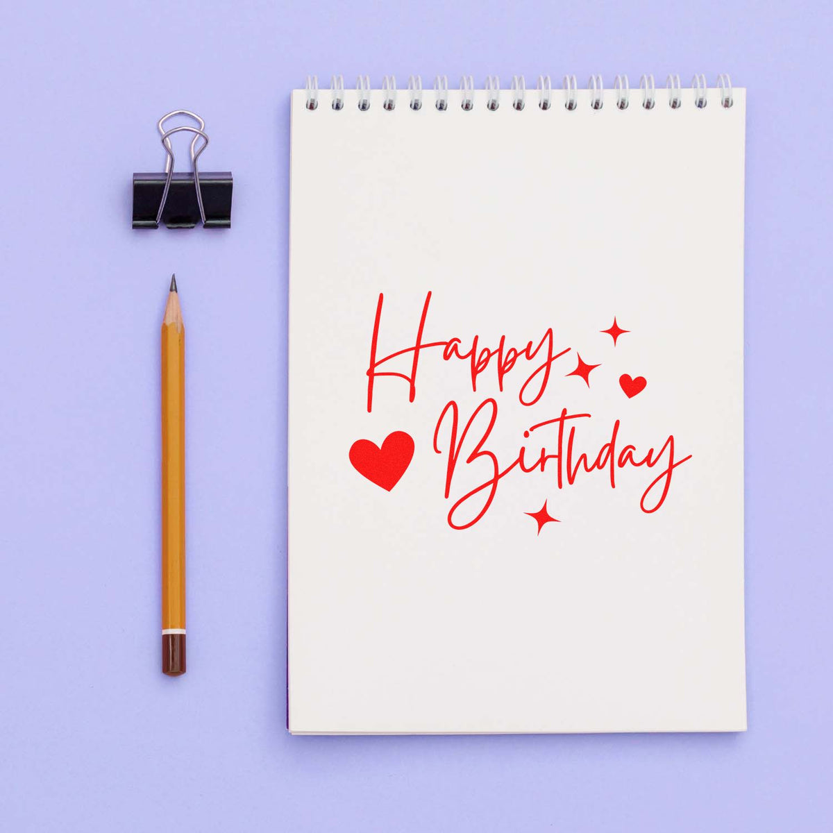 Red Happy Birthday stamp/seal on white spiral notebook with hearts and sparkles, beside a black binder clip and yellow pencil on lavender background.