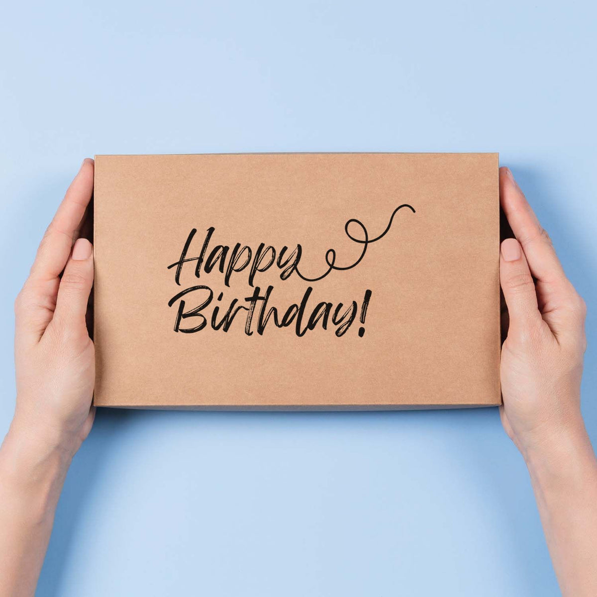 Hands holding a kraft box with black Happy Birthday stamp on a light blue background.