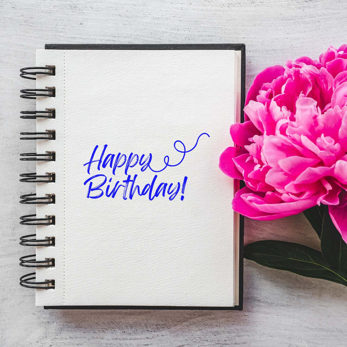 Blue Happy Birthday stamp on a white spiral notebook, with bright pink peonies and green leaves.