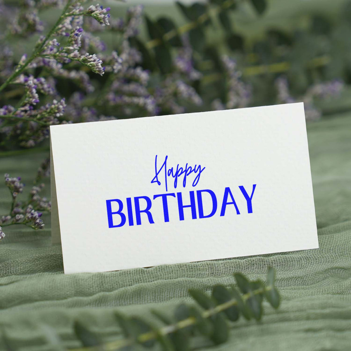 Blue Happy Birthday stamp on a white card, set among green foliage and purple flowers for a festive seal product.