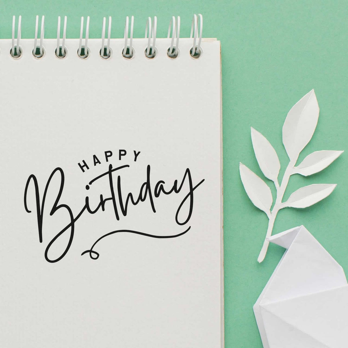 Happy Birthday stamp and seal on white spiral notebook, with mint green background, white paper leaves and origami gift.
