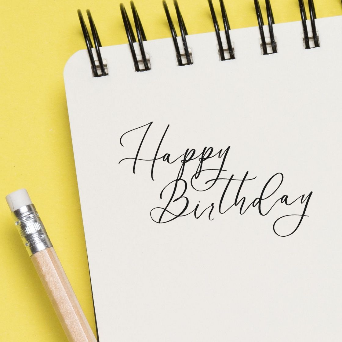 Happy Birthday in black script on a white spiral notebook, with a pencil on a yellow background; stamp and seal product.