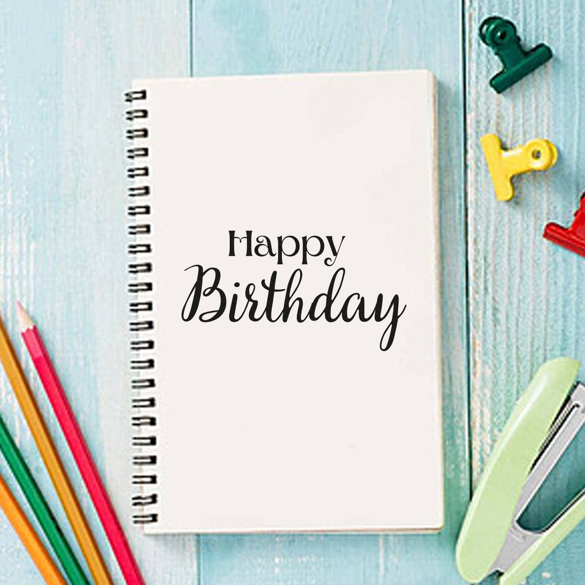 Happy Birthday notebook with black script, colorful stamp and seal clips, and pencils on blue wood background.