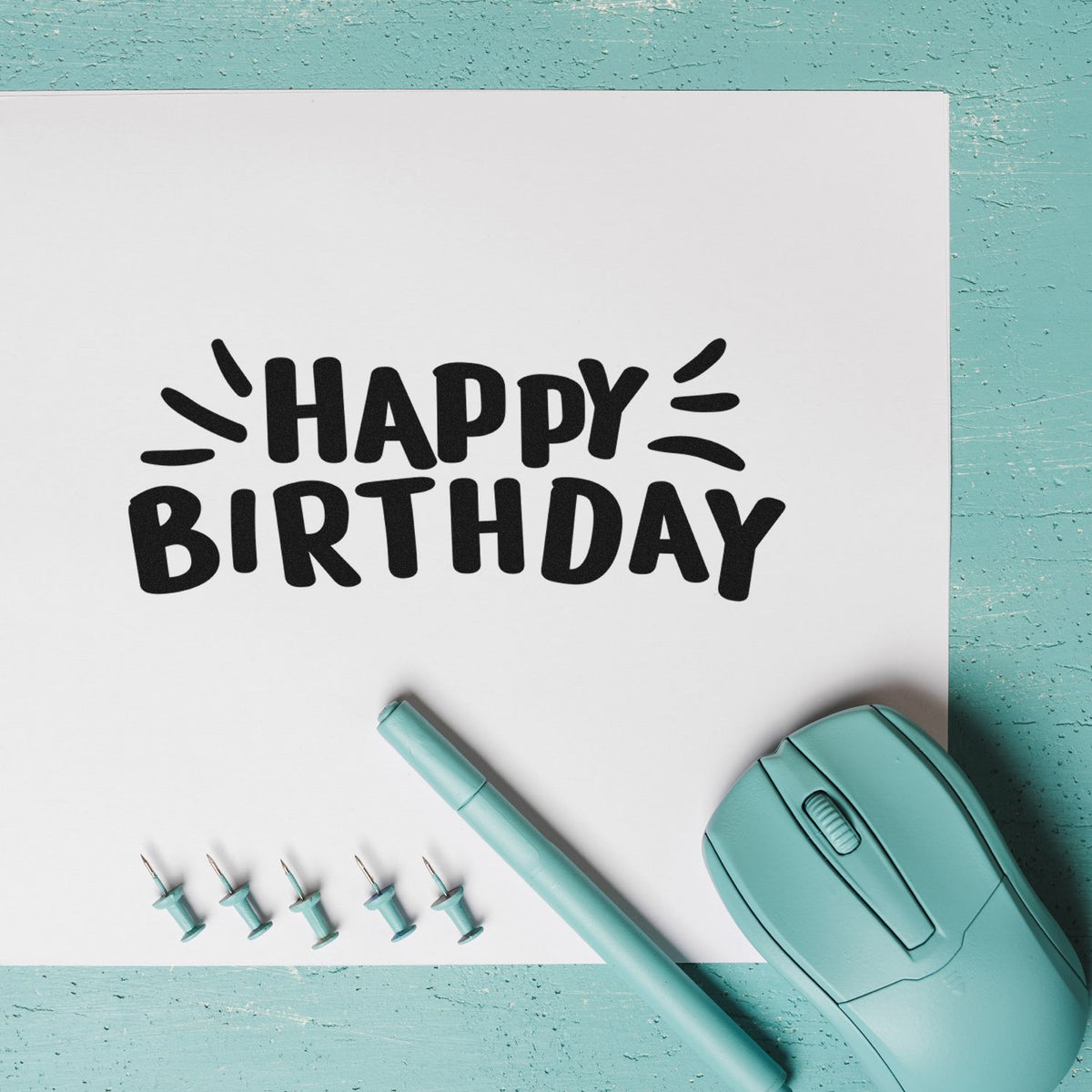 Black Happy Birthday stamp design with teal stamp seal tool, pen, and pins on white paper over aqua background.