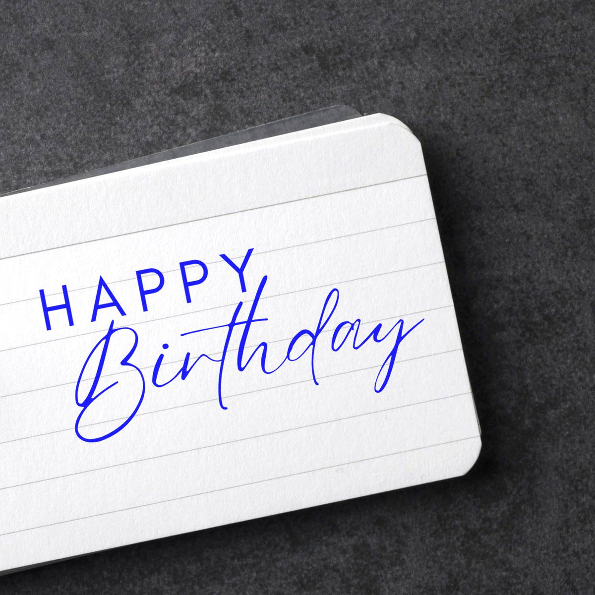 Blue Happy Birthday stamp and seal on white lined card against a dark textured background.