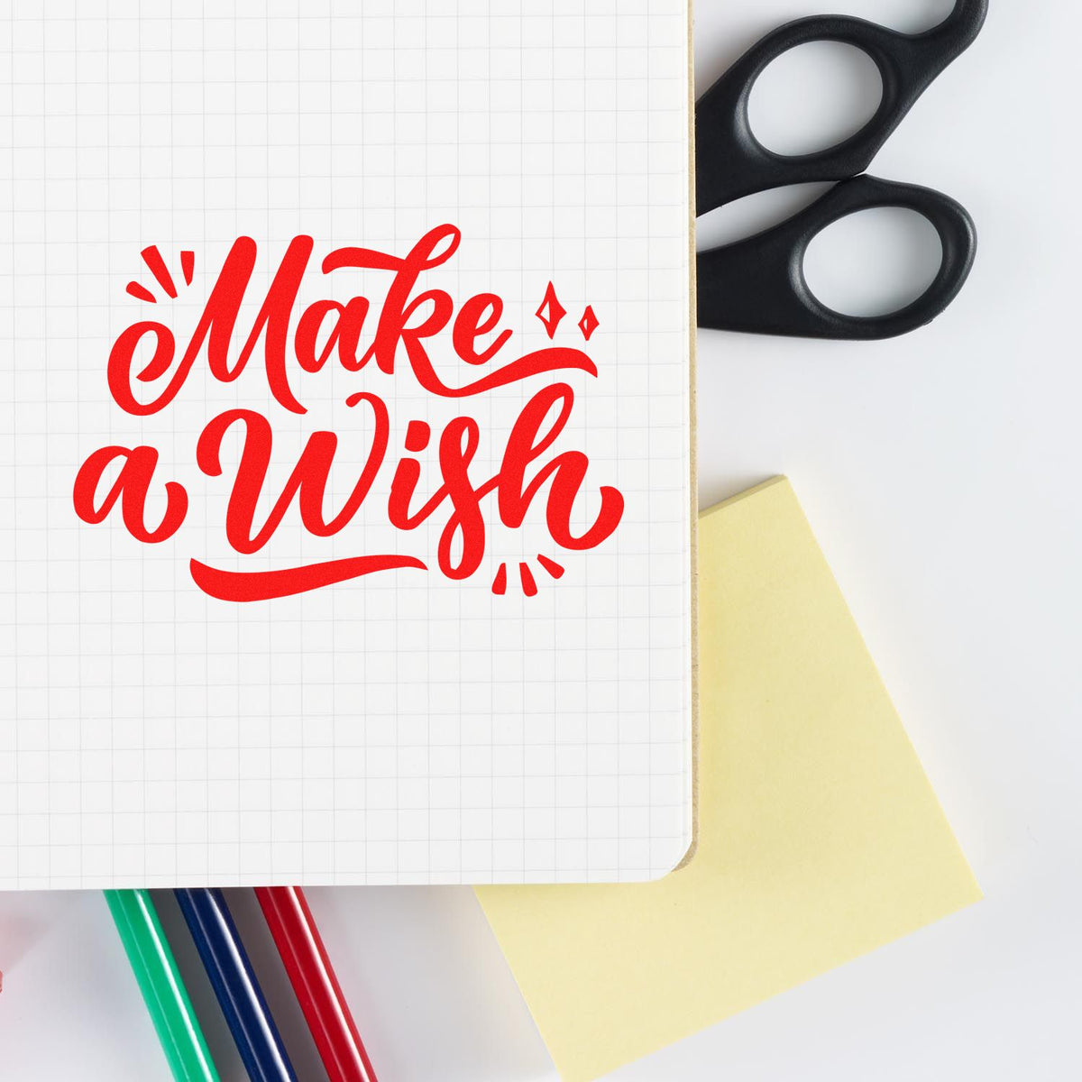 Red Make a Wish stamp on a graph notebook, with black scissors, yellow sticky note, and colored pens on white background.