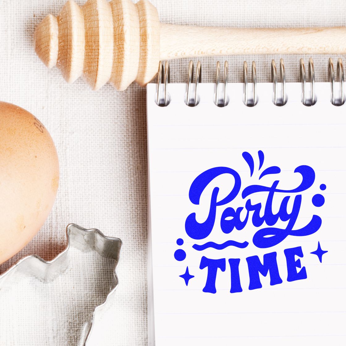Blue Party Time stamp and seal design on a spiral notepad, with wooden honey dipper, egg, and metal cutter on beige fabric.