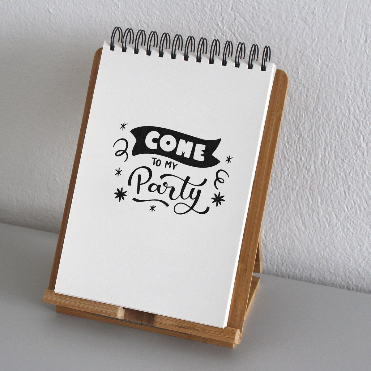 White spiral notepad on a wooden stand with black Come to My Party stamp seal and star accents.