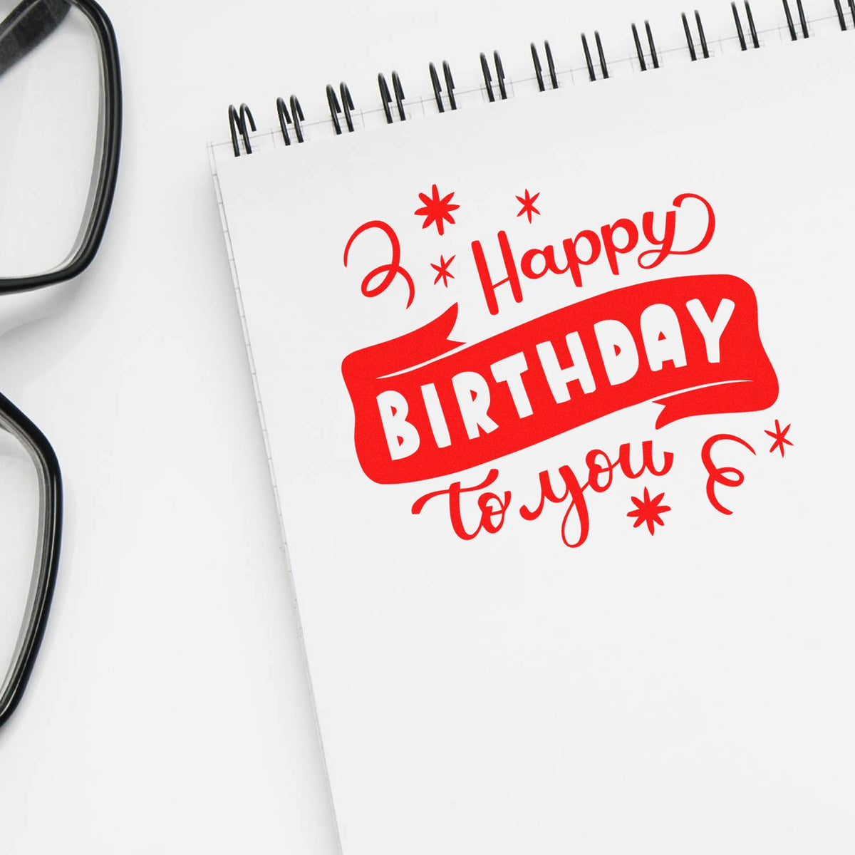Red Happy Birthday stamp on a white spiral notebook, with black eyeglasses at the left edge.