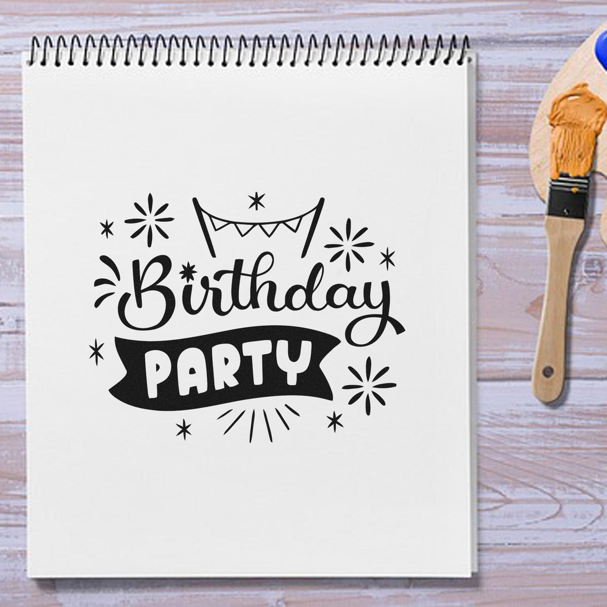 Black birthday party stamp design on white spiral notebook, with orange inked seal brush on a light wood surface.