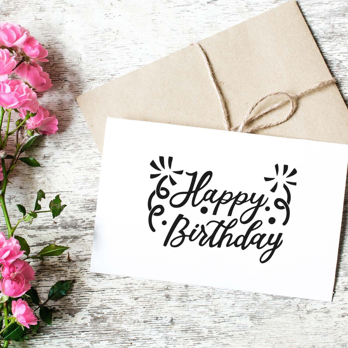 Black Happy Birthday stamp and seal design on white card, with beige twine-wrapped envelope and pink flowers on wood.