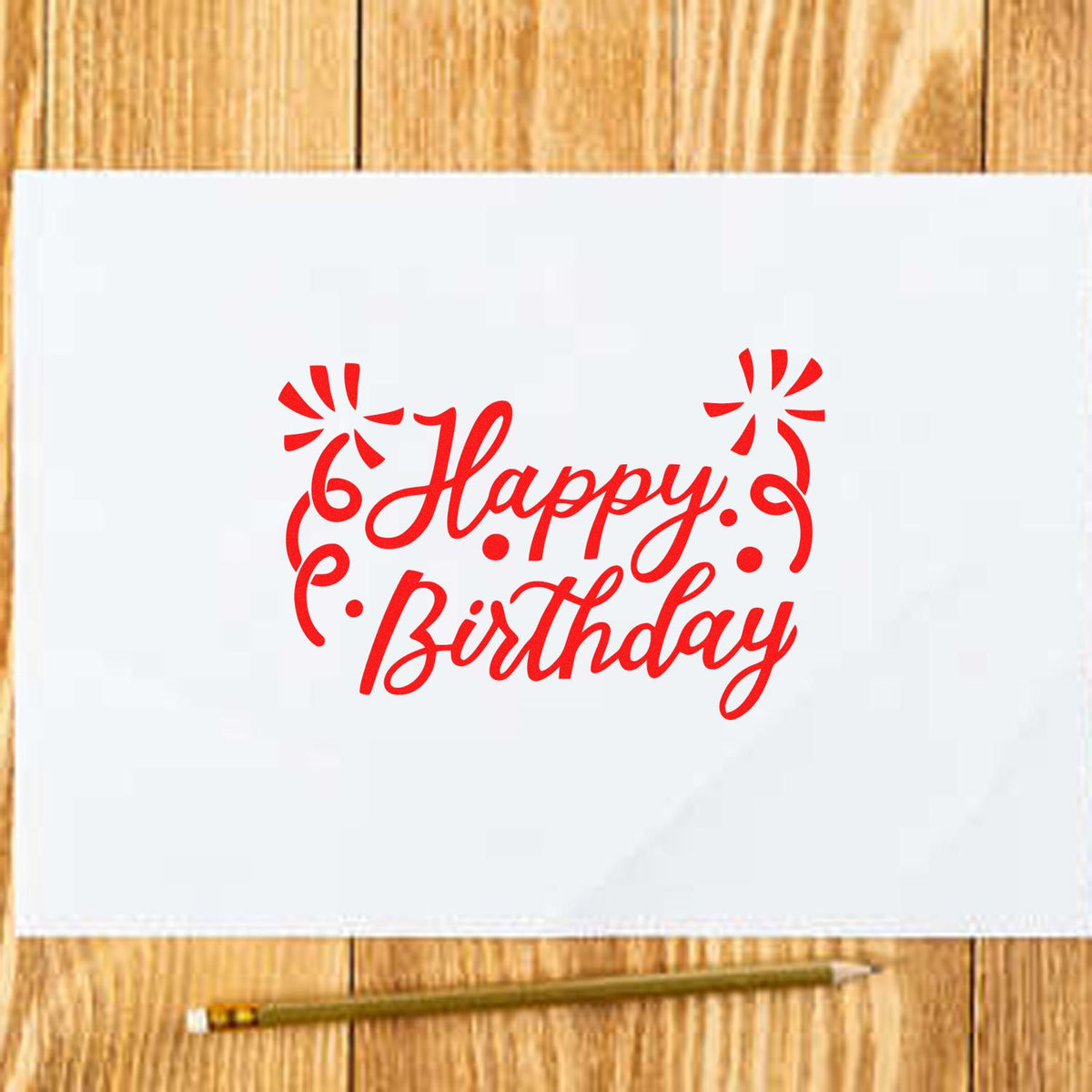 Red Happy Birthday stamp/seal design on white paper, with decorative swirls, on a wooden table beside a pencil.