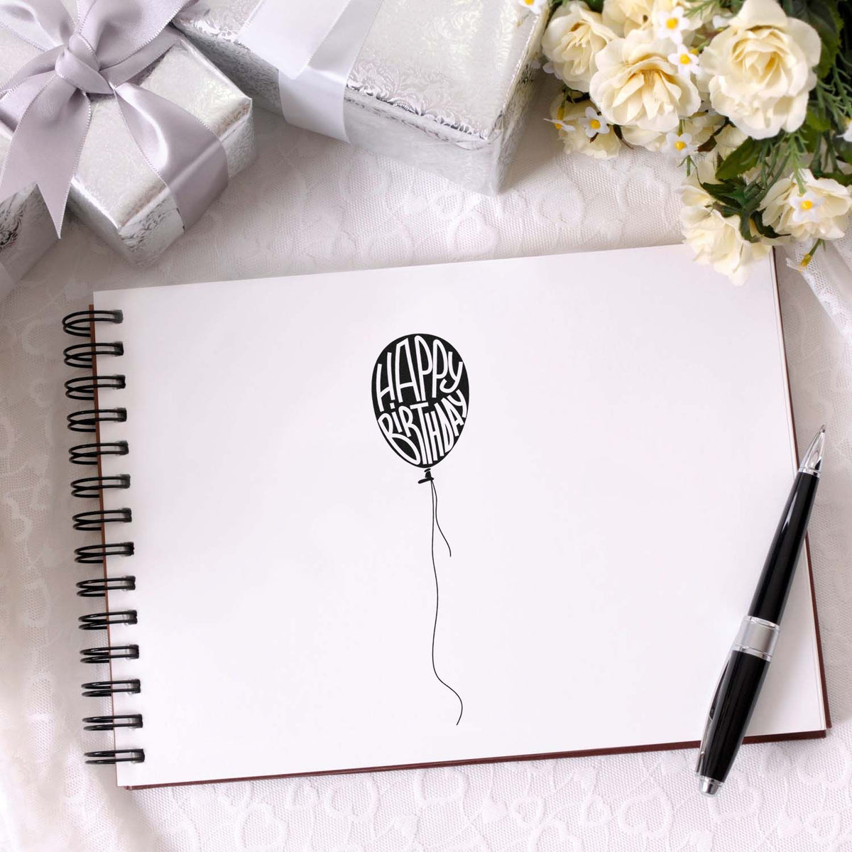 Black Happy Birthday balloon stamp and seal on white spiral notebook, with silver gift boxes, black pen, and cream flowers.