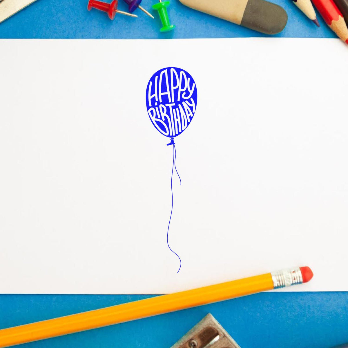 Blue Happy Birthday balloon stamp seal on white paper, framed by pencils, push pins, and a yellow pencil.